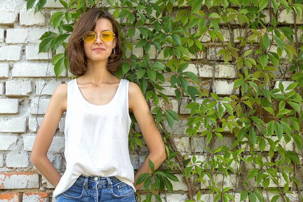 A woman dressed in a summer layering style. She wears a white linen tank top, an oversized sheer button-down shirt, denim shorts, and sandals. She is accessorized with a wide-brimmed straw hat and sunglasses.