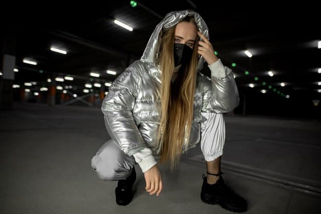 A street style shot of a woman wearing an oversized puffer jacket in a metallic silver color. She is paired with black leggings, chunky sneakers, and sunglasses. The backdrop shows a bustling city street.