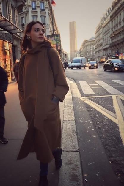 A French woman is walking down a street in Paris, wearing a long beige trench coat, a white scarf, and sunglasses. She is carrying a leather handbag and wearing ankle boots. The background shows classic Parisian buildings and street signs.